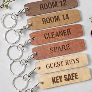 May include: A set of six rectangular wooden keychains with silver metal rings and clips. Each keychain features engraved text: ROOM 12, ROOM 14, CLEANER, SPARE, GUEST KEYS, and KEY SAFE. The wood tones vary from dark brown to light tan, showcasing natural grain patterns.