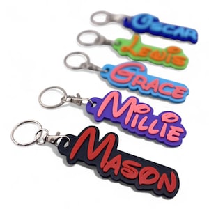 May include: A collection of colorful name keychains. Each keychain features a different name: Oscar, Lewis, Grace, Millie, and Mason. The names are in various colors, with a 3D effect, and attached to a silver keyring.