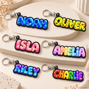 Peut inclure: Six porte-clés personnalisés avec des fermoirs mousquetons argentés. Les prénoms Noah, Oliver, Isla, Amelia, Riley et Charlie sont inscrits en lettres style bulle colorées avec des motifs variés incluant des étoiles, des cœurs et des dinosaures.