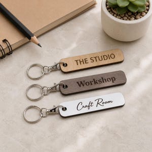 May include: Three rectangular keychains with silver metal rings and clips. The top keychain is light wood with the text THE STUDIO, the middle is dark wood with the text Workshop, and the bottom is white with the text Craft Room in script.