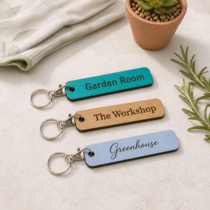 May include: Three rectangular keychains with metal rings. The top one is teal with the text Garden Room. The middle one is natural wood with the text The Workshop. The bottom one is light blue with the text Greenhouse in cursive script.