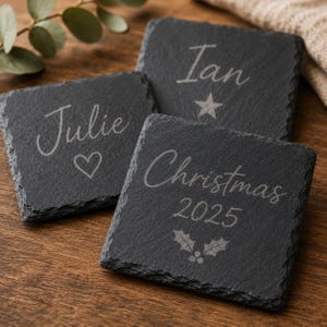 Puede incluir: Tres posavasos cuadrados de pizarra negra con bordes naturales y rugosos. Cada posavasos est&aacute; grabado con texto y s&iacute;mbolos en blanco: Julie con un coraz&oacute;n, Ian con una estrella y Christmas 2025 con un dise&ntilde;o de hojas de acebo y bayas.
