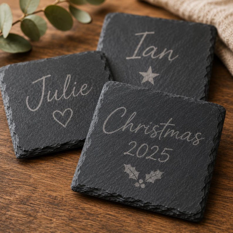 Puede incluir: Tres posavasos cuadrados de pizarra negra con bordes naturales y rugosos. Cada posavasos est&aacute; grabado con texto y s&iacute;mbolos en blanco: Julie con un coraz&oacute;n, Ian con una estrella y Christmas 2025 con un dise&ntilde;o de hojas de acebo y bayas.