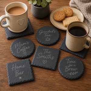 Puede incluir: Una colecci&oacute;n de posavasos de pizarra gris oscuro en formas cuadradas y circulares, con textos grabados que incluyen Grandad's Tea, Mum's Coffee, The Smiths, Home Bar y Time for a Brew, junto a dos tazas de cer&aacute;mica y un plato de galletas.