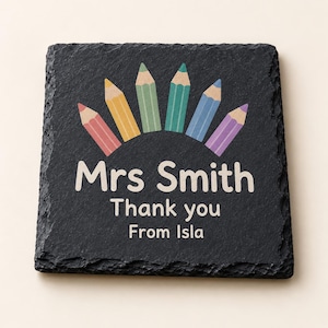May include: A square black slate coaster with a rough, textured edge. The center features a graphic of six colored pencils arranged in a semi-circle above the white printed text: Mrs Smith, Thank you, From Isla. The design is centered on the stone.