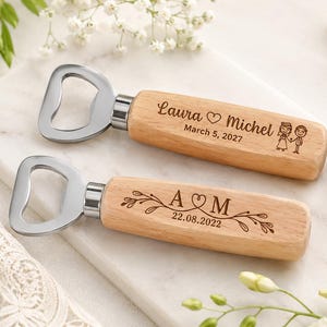 May include: Two bottle openers with light-coloured wooden handles and silver metal heads. The top opener is engraved with "Laura ♡ Michel", "March 5, 2027", and a cartoon couple. The bottom opener features "A ♡ M", "22.08.2022", and leaf designs.