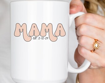 May include: White ceramic mug with the word "MAMA" in a retro font, outlined in brown, and the text "EST. 2-12-24" below. The mug has a large handle and is held by a person. A gold ring is visible on the person's finger.