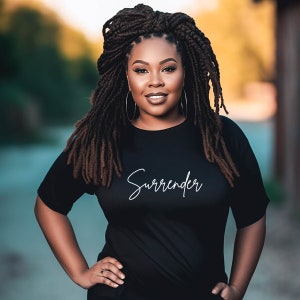 May include: A woman wearing a black t-shirt with the word "Surrender" in white script. She has long, dark dreadlocks and large gold hoop earrings. She is smiling and has her hands on her hips.