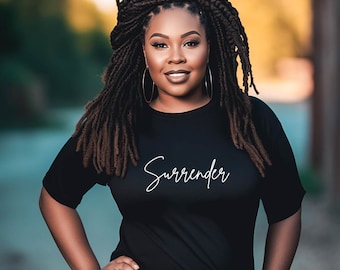 Peut inclure: Une personne portant un t-shirt noir avec le mot "Surrender" en écriture blanche. Elle a de longues dreadlocks foncées et de grandes créoles dorées. Elle sourit, les mains sur les hanches.