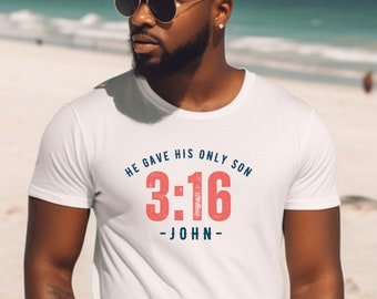 Puede incluir: Camiseta blanca con el texto "HE GAVE HIS ONLY SON 3:16 - JOHN" en azul y rojo. La camiseta es de cuello redondo. La imagen está tomada al aire libre con una playa y el océano de fondo.