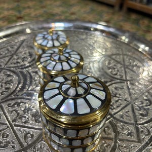 Könnte beinhalten: Drei dekorative Schmuckdosen mit Perlmutt-Inlay und goldfarbenen Verzierungen. Die Dosen sind rund mit gewölbtem Deckel und stehen auf einem silbernen Tablett. Das Inlay erzeugt ein Mosaikmuster mit schwarzen Akzenten.