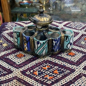 May include: A ceramic spice holder with six compartments, featuring a central lid and brass accents. The holder is painted with blue, green, and white patterns. It sits on a patterned purple and white textile.