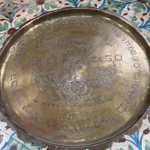 May include: A round brass tray with a scalloped edge featuring intricate Hebrew engravings and floral motifs. The centre displays decorative script and symmetrical patterns, while the outer rim is bordered by a continuous band of Hebrew text.