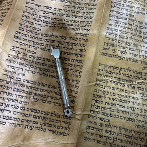 Może przedstawiać: Srebrny wskaźnik z gwiazdkowym zakończeniem spoczywa na pergaminowym zwoju z hebrajskim tekstem. Zwój jest postarzany, o ciepłym, żółto-brązowym odcieniu, a tekst jest czarny.