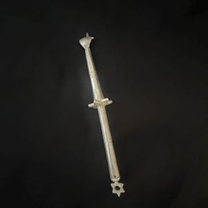 Könnte beinhalten: Ein silberfarbenes, längliches Objekt mit dekorativen Ätzungen und stern- und vogelförmigen Endstücken. Das Objekt hat ein zentrales, kreisförmiges Design und ist vor einem schwarzen Hintergrund platziert. Das Objekt scheint ein dekorativer Gegenstand zu sein.