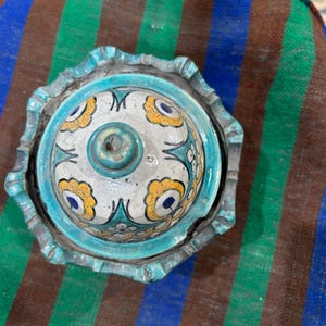 Puede incluir: Un plato de cerámica para baratijas con tapa. El plato y la tapa son turquesas con un diseño floral en amarillo y azul. El plato se encuentra sobre un textil a rayas marrón, verde y azul.