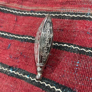 May include: A silver-colored, elongated bottle stopper with intricate floral patterns etched on its surface. The stopper has a pointed top and a decorative base. It rests on a red and black striped textile.