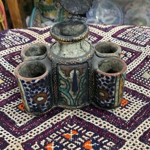 以下が含まれることがあります： ヒンジ付きの蓋と4つの小さなカップのようなホルダーが付いた、アンティークで装飾的な金属製の容器。容器は、青、オレンジ、緑の複雑な模様で、風化した、古びた外観をしています。模様のある紫と白の織物の上に置かれています。
