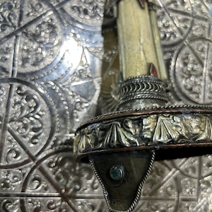 Pode incluir: Close-up de um castiçal prateado ornamentado com uma vela creme. O castiçal apresenta intrincados trabalhos em metal, incluindo desenhos florais e uma pequena pedra turquesa. O fundo é uma superfície prateada padronizada.