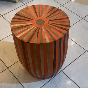 Peut inclure: Un tabouret rond en bois avec un motif rayé orange, marron et blanc. Le dessus du tabouret a un motif circulaire au centre.