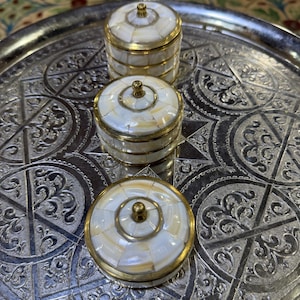 Könnte beinhalten: Drei kleine, runde Behälter mit Deckel, die aus einem Mosaik aus weißen und cremefarbenen Muschelsegmenten in Messingrahmen bestehen. Jeder Behälter hat einen kleinen, mittigen Messingknopf und steht auf einem runden, silberfarbenen Metalltablett.