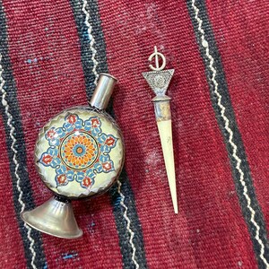 May include: A round metal bottle with a narrow neck and a flared base, featuring a central orange, blue, and red floral pattern. Beside it lies a long, pointed applicator tool with a triangular silver top and a thin, off-white tapered end.