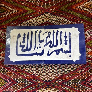 Peut inclure: Deux carreaux rectangulaires avec calligraphie arabe bleue sur fond blanc, encadrés d'une bordure bleue. Les carreaux sont posés sur un tapis coloré à motifs aux teintes rouges, vertes et oranges.