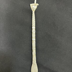 May include: A silver-coloured, long, slender utensil with intricate geometric patterns. The top features a hand-shaped design with a raised index finger. The handle is adorned with detailed carvings, and the bottom has a rounded, decorative end.