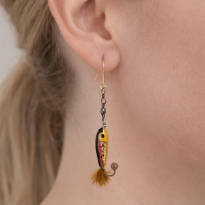 May include: A single gold-coloured earring with a miniature fishing lure design. The lure is yellow, black, and red, with a small brown feather and a hook. The earring hangs from a gold-coloured ear wire.