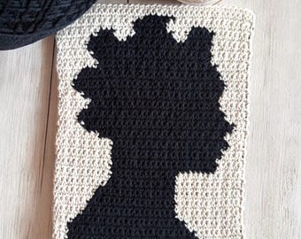 May include: A rectangular crochet piece featuring a black silhouette of a person with a textured afro hairstyle centered against a cream-colored background. The design is created using a tapestry crochet technique with visible yarn stitches throughout.