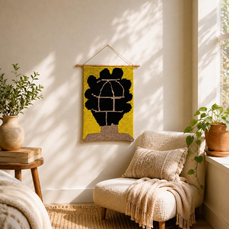Puede incluir: Un tapiz rectangular con fondo amarillo y una silueta negra abstracta de cabeza y hombros. El dise&ntilde;o incluye un dibujo lineal blanco de una jaula de p&aacute;jaros dentro de la forma negra. Cuelga de un soporte de madera con una cuerda.