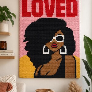 May include: A rectangular crochet wall hanging featuring the word LOVED in bold red letters at the top. The design depicts a person with dark skin, voluminous black hair, white-rimmed sunglasses, and large white hoop earrings against a pink background.