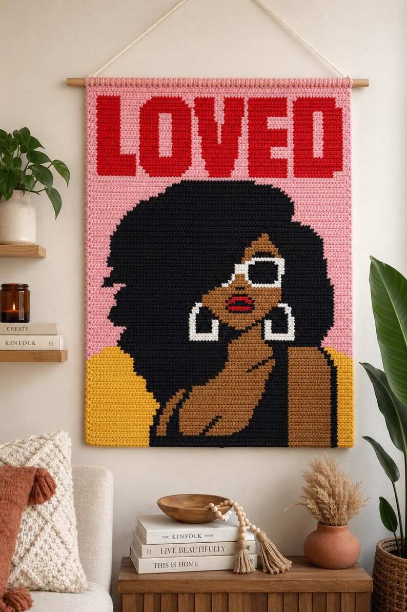 May include: A rectangular crochet wall hanging featuring the word LOVED in bold red letters at the top. The design depicts a person with dark skin, voluminous black hair, white-rimmed sunglasses, and large white hoop earrings against a pink background.