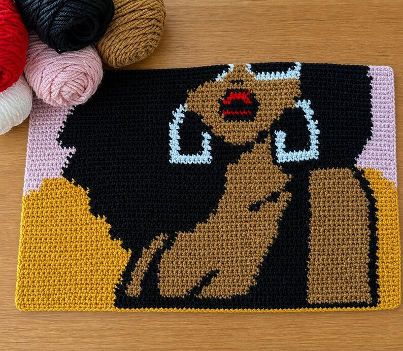 May include: A rectangular crochet tapestry featuring a graphic portrait of a person with dark skin, voluminous black hair, and large light blue hoop earrings. The figure has bright red lips and is set against a background of mustard yellow and light pink.
