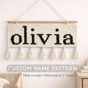 May include: A rectangular cream-coloured crochet wall hanging featuring the name olivia in black lowercase letters. The piece is suspended from a light brown wooden dowel with a white string and includes six cream-coloured tassels along the bottom edge.