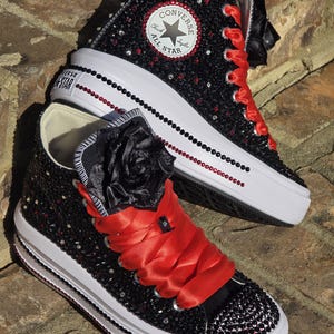May include: Black high-top Converse sneakers with red ribbon laces and a black flower embellishment. The sneakers are covered in black rhinestones and have a white rubber sole. The Converse logo is visible on the side of the shoe.