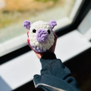 May include: A white and purple crocheted stuffed animal with a pig-like face. The animal is being held by a hand.
