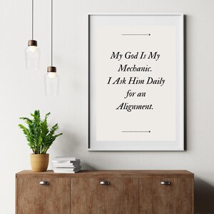 Puede incluir: Una impresión enmarcada con el texto "My God Is My Mechanic. I Ask Him Daily for an Alignment." La impresión está en un marco blanco y cuelga sobre un mueble de madera con una planta y luces decorativas.