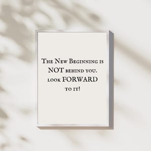 Peut inclure: Une affiche encadrée avec le texte "The New Beginning is NOT behind you. Look forward to it!" en noir sur fond crème. Le cadre est argenté et le fond est beige clair.