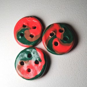 May include: Three round buttons featuring a marbled swirl pattern of red, dark green, and brown. Each button has a smooth, glossy finish and contains four holes arranged in a square pattern for attachment to fabric or other materials.