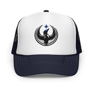 Könnte beinhalten: Trucker-Cap mit weißem Frontpanel und marineblauem Schirm und Mesh-Rückseite. Die Kappe hat ein schwarz-blaues gesticktes Design eines Vogels mit ausgebreiteten Flügeln und einem Stern darüber. Die Kappe hat einen gebogenen Schirm.