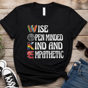Peut inclure: Un t-shirt noir à manches courtes avec le texte blanc WOKE, où le W est un motif de crayons colorés, le O un symbole de poing noir, le K une lettre arc-en-ciel et le E un symbole féminin rouge, suivi de ISE, PEN MINDED, IND AND et MPATHETIC.