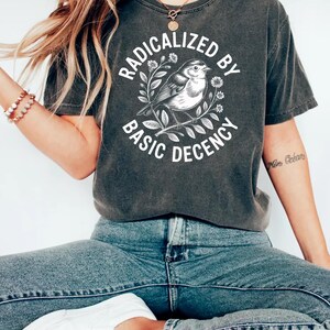Puede incluir: Camiseta gris oscuro con el texto "RADICALIZED BY BASIC DECENCY" en un diseño circular alrededor de una ilustración de un pájaro. La camiseta se combina con vaqueros azules y zapatillas blancas.