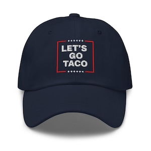 May include: A navy blue baseball cap with the embroidered text "LET'S GO TACO" in white, framed by a red rectangle and small white stars. The cap has a curved brim and a top button.
