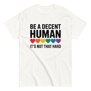 Könnte beinhalten: Ein weißes kurzärmeliges T-Shirt mit dem schwarzen Text BE A DECENT HUMAN IT'S NOT THAT HARD. Zwischen den beiden Textzeilen befindet sich eine horizontale Reihe aus sechs kleinen Herzen in den Farben Rot, Orange, Gelb, Grün, Blau und Lila.