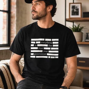 Może przedstawiać: Czarny t-shirt z krótkim rękawem z białym nadrukiem graficznym w formie poziomych pasków oraz tekstem "everything is fine trust your government" pośrodku. Koszulka ma okrągły dekolt i standardowy krój, wykonana z jednolitego czarnego materiału.