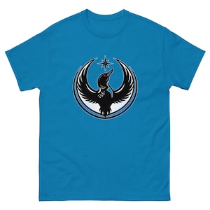 May include: A turquoise blue t-shirt featuring a black and white graphic of a bird with outstretched wings inside a circular frame. The bird has a red eye and is positioned below a white star.