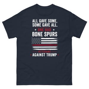 以下が含まれることがあります： 白文字とアメリカ国旗のデザインが施された、濃いネイビーブルーのTシャツ。「ALL GAVE SOME, SOME GAVE ALL. ONE HAD BONE SPURS. VETERANS AGAINST TRUMP.」と書かれています。