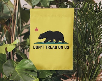 May include: Yellow garden flag featuring a black bear silhouette, a red star, and the text "DON'T TREAD ON US." The flag is displayed on a black metal stand, surrounded by green plants.
