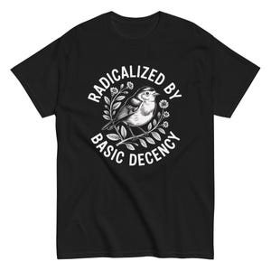 Puede incluir: Camiseta negra con un diseño gráfico blanco de un pájaro posado en una rama, rodeado de flores y el texto "RADICALIZED BY BASIC DECENCY" en un diseño circular.
